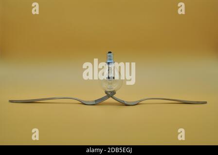 light bulb balancing on composition of 2 intertwined forks isolated on orange neutral background, conceptual photo to indicate division of electricity Stock Photo