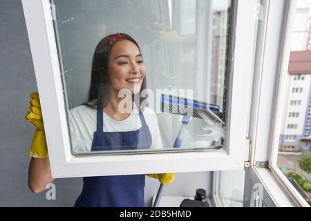 Fast and efficient. Attractive young woman smiling while cleaning the window using squeegee. Housework and housekeeping, cleaning service concept Stock Photo