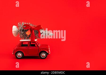 Red toy car carrying Christmas tree with red ribbon on top of the roof on red background. Space for text on the right. Stock Photo