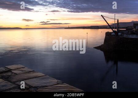 Morning light in Stromness bay with stone slabs in foreground and sea and lighthouse in the background Stock Photo