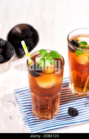 The glass of iced tea with blackberry on wooden table. Cuba Libre or long island cocktail, cold drink or lemonade with lemon and basil. Stock Photo