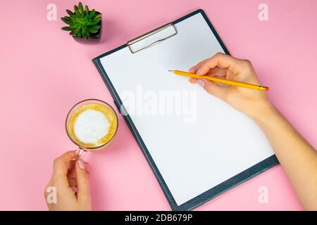 Tablet with white flattery next to which stands a green plant, hand holds a pencil and draws on a sheet in the other hand a cup with a coffe. Stock Photo