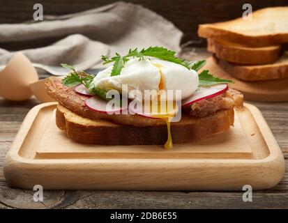 sandwich on toasted white slice of bread with poached eggs, green leaves of arugula and radish, morning breakfast on a brown wooden board Stock Photo