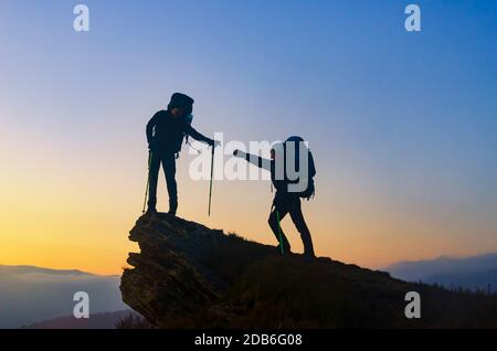 Helping hand on the top. Hiker with backpack helps other hiker to reach the summit. Concept of help, achievement and friendship. Stock Photo