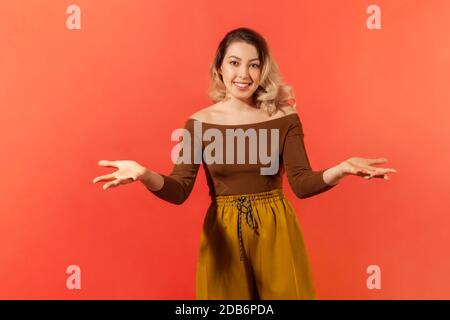 Portrait of happy young woman with blonde hair in brown blouse standing and opening her hands with ready to hug, offer for share or take for free. Ind Stock Photo