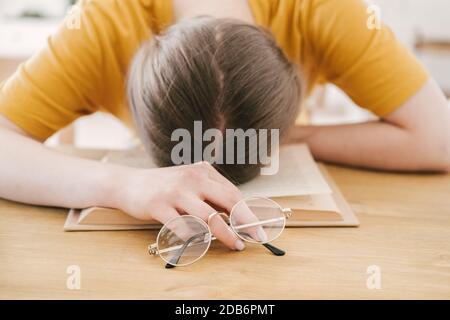 Young beautiful woman student in orange T-shirt and glasses reads book sitting at the table. Preparing for exams, studying at home on self-isolation Stock Photo