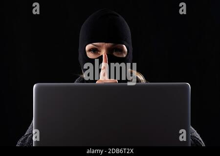 Cyber terrorist in a mask in a mask on a black background behind a laptop Stock Photo