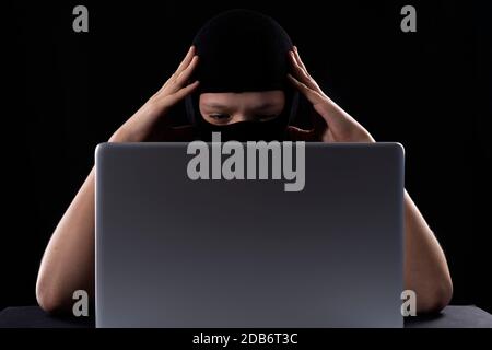 Cyber terrorist in a mask in a mask on a black background behind a laptop Stock Photo