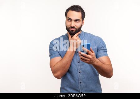 Thoughtful bearded man scratching chin holding smartphone in hand, contemplating about software updating, choosing suitable tariffs. Indoor studio sho Stock Photo