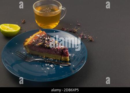 Slices of blueberry mousse cake with mirror glaze decorated berries and leaves of mint on a blue plate with a mug of mint tea and lemon are on black t Stock Photo