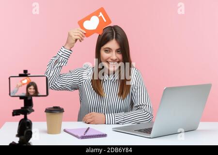 Smiling positive woman blogger in striped shirt recording video for followers holding like sign above head, asking to rate her posts and subscribe. In Stock Photo