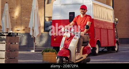 Young handsome caucasian man courier in red uniform driving scooter, smiling at camera. Food delivery concept Stock Photo