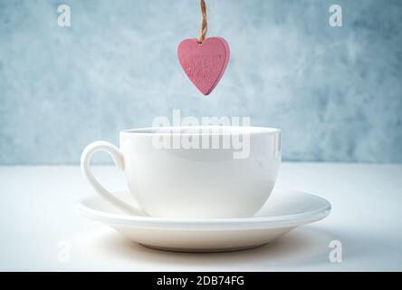 A pink heart hangs over a coffee Cup and saucer on a white and gray background. Stock Photo