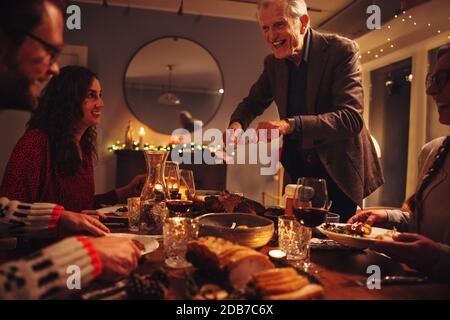 Elderly man serving food to his family sitting at dinner table on Christmas eve. Family having Christmas dinner together in warn and cozy scandinavian Stock Photo