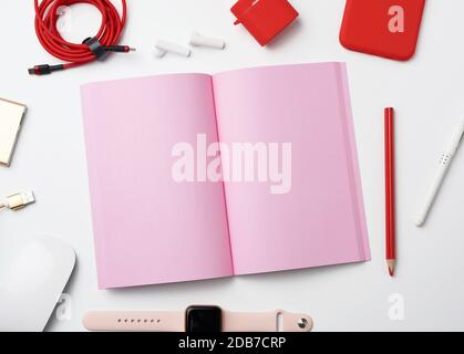 pink paper notebook, power bank with cable, red smartphone, headphones, wireless mouse, smart watch on white background, workplace, top view Stock Photo
