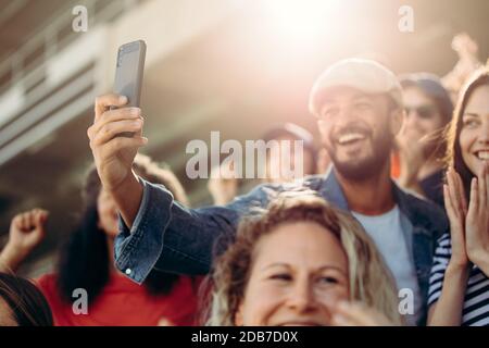 Young man and woman taking selfie at stadium with spectators around. Excited couple taking selfie while watching match in stadium. Stock Photo