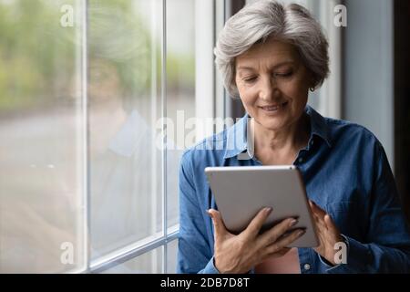 Close up smiling mature woman using tablet, standing near window Stock Photo