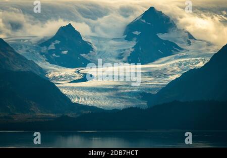 The morning sun rises over Kachemak Bay near Homer Alaska with clouds rolling over the mountain peaks and glaciers Stock Photo