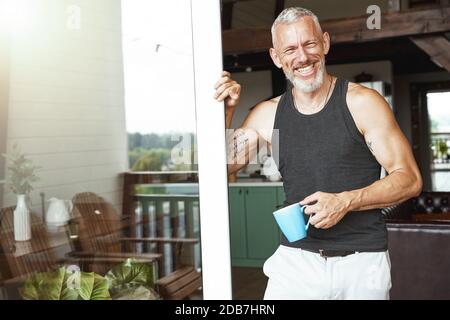 Perfect morning. Handsome positive middle aged caucasian man with coffee standing near terrace entrance of his modern apartment Stock Photo