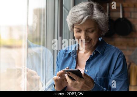 Close up smiling mature woman using smartphone, standing near window Stock Photo