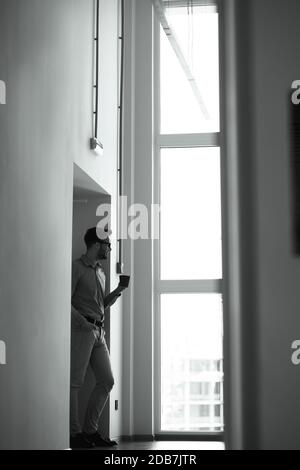 Vertical shot of a young businessman drinking fresh coffee while standing in the office and looking out the window, black black and white photo Stock Photo
