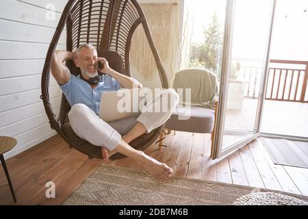 Perfect workplace. Happy middle aged caucasian businessman talking by phone while sitting with laptop on swing chair at his modern apartment, working Stock Photo