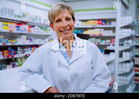 Smiling senior pharmacist looking happy leaning against medication counter standing in pharmacy  Stock Photo