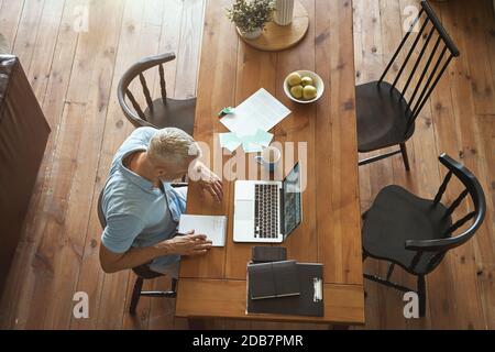Top view of a mature caucasian businessman making some notes and using laptop while sitting at table in his modern apartment Stock Photo