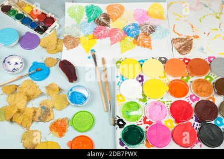 Finger paints and watercolors on the child table. Painted autumn tree leaves imprinted on the paper.  Toys for encouraging creativity of children. Stock Photo
