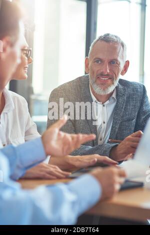 Cheerful mature businessman sitting with colleagues in the modern office and discussing something Stock Photo