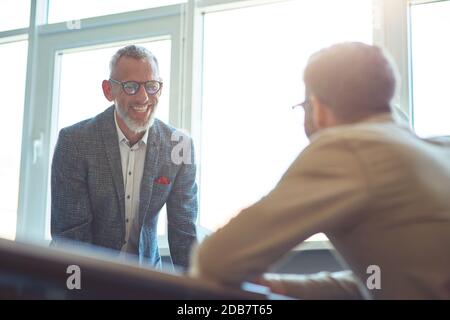 Mature cheerful businessman discussing something with his young male colleague and smiling, working together in the modern office Stock Photo