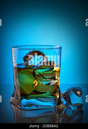 Whiskey in transparent glass with ice cube lying on glass table on blue background Stock Photo