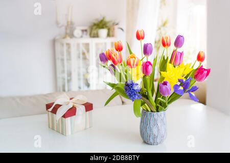 Fresh spring colorful bouquet of tulips, daffodils, irises in vase and gift box on white table with light classic design room background. Festive Stock Photo