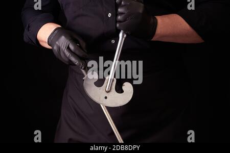 male cook in black uniform and black latex gloves holds a large sharp vintage knife for meat and vegetables and a metal sharpener for knives, black ba Stock Photo