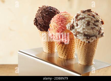Three ice cream cones with assorted flavors including chocolate, strawberry and choc chip standing in a metal holder on a counter