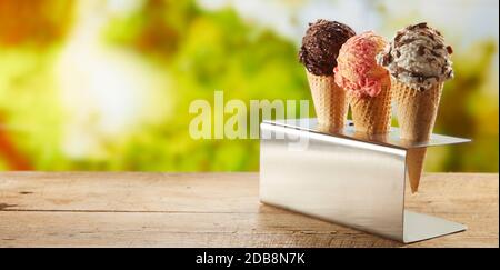 Three flavors of ice cream in cones on an outdoor table standing in a metal holder with blurred garden backdrop of green trees and copy space in panor Stock Photo