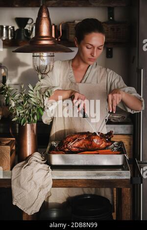 Young woman cooking and carving whole oven roasted duck for Thanksgiving holiday or Christmas party celebration in kitchen interior. Traditional Autumn or Winter holiday comfort food for gathering Stock Photo