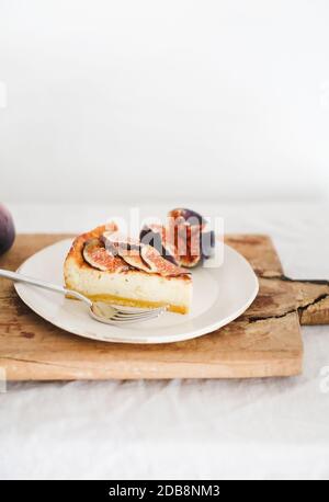 Slice of homemade gluten free fig cheesecake with fresh seasonal figs on white plate over rustic wooden board, white wall at background. Healthy dessert, artisan sweet food concept Stock Photo