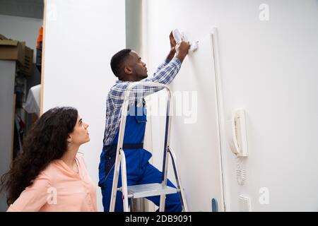 Happy Woman Looking At Electrician Installing Security System Motion Detector On Wall Stock Photo