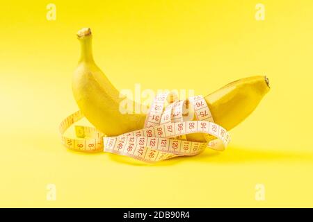 Banana with tape for measuring figure. Centimeter ruler spinned around fruit. Tape wrapped around banana isolated on yellow background. Weight loss, h Stock Photo