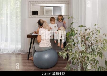 Mother sits on a fitness ball in headphones and works at home with her children during quarantine Stock Photo