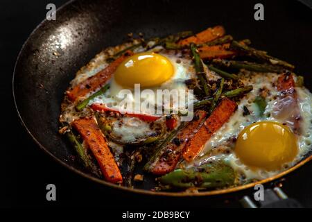 Very coarsely two fried eggs in a pan with large slices of carrots, bell peppers and asparagus with spices and sauce. Stock Photo