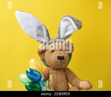 cute brown teddy bear holding colorful Easter eggs, wearing a rabbit mask with long ears on his head, funny holiday card Stock Photo