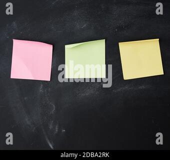 three blank paper multi-colored square stickers are stuck on a black chalk board Stock Photo