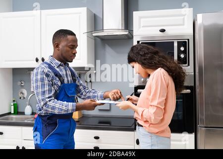 Smiling Customer Woman Signing Appliance Repair Invoice In Front Of Plumber Standing In Kitchen Stock Photo