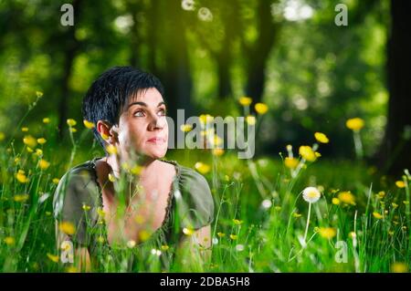 Low angle view of a middle-aged woman lying to one side on her stomach in the middle of a meadow littered with yellow flowers on the edge of the fores Stock Photo