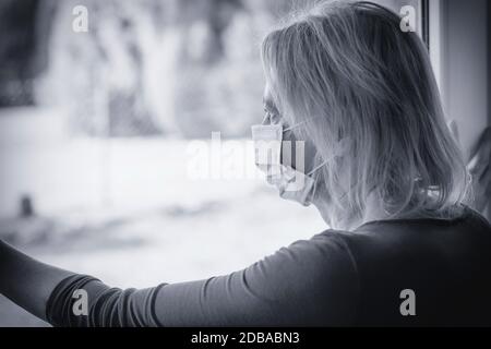 Lonely woman in a quarantine with a disposable protective face mask looks sadly out of the window. Stock Photo