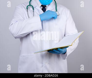 doctor in a white coat with buttons, wearing blue sterile gloves, holding a paper open notebook in his hand on a white studio background Stock Photo