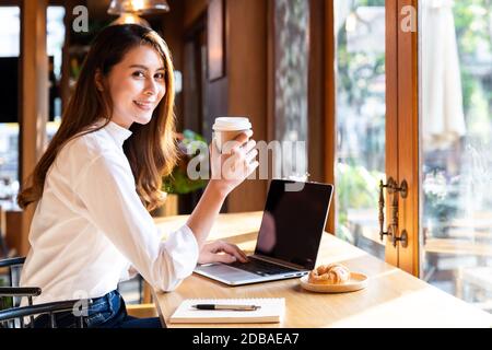 Quarantine Young asian woman wearing smart casual clothes work at home in living room using laptop and drinking hot coffee and croissant while city  l Stock Photo