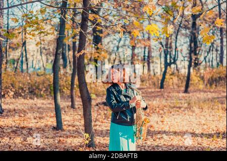 pretty dark-haired girl in a black jacket and blue skirt with a saxophone in a yellow autumn park Stock Photo